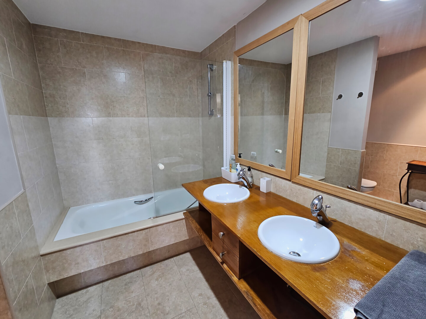 Baño común con dos lavabos sobre mueble de madera, bañera integrada con mampara, espejo grande y acabados de calidad