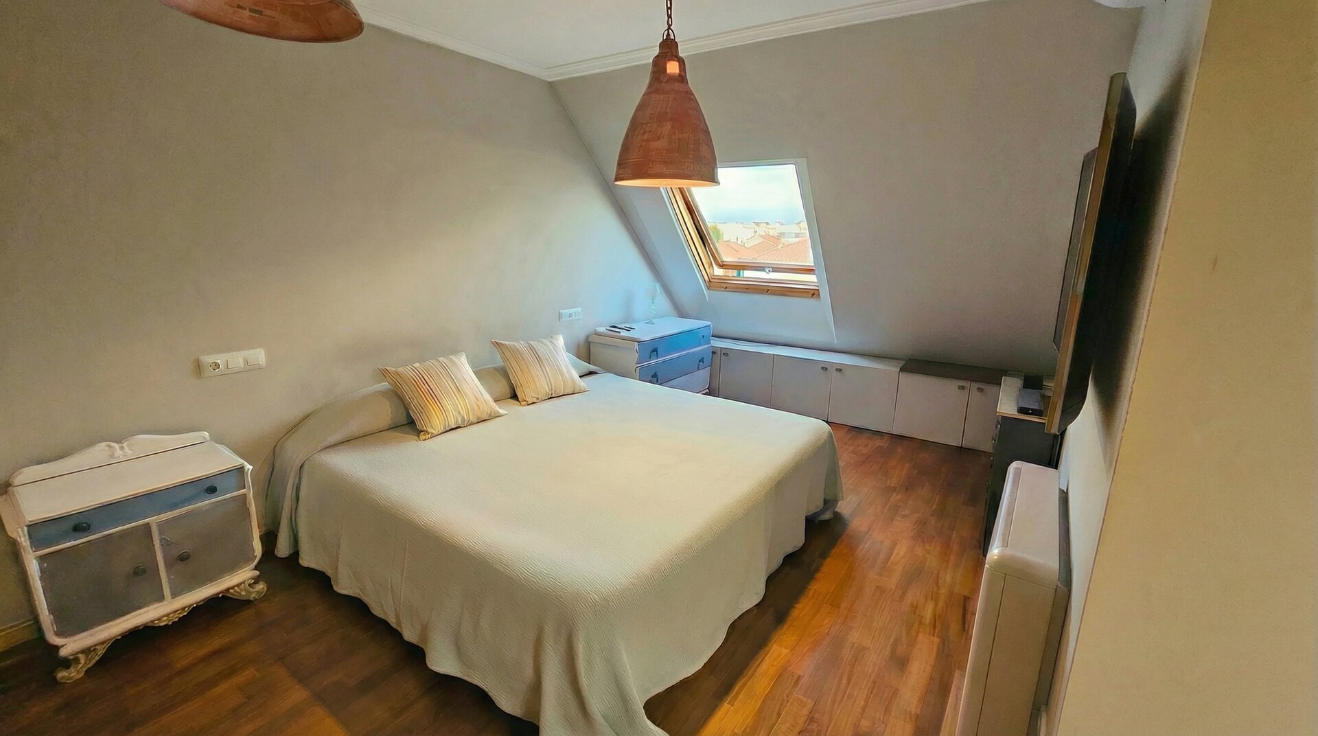 Dormitorio principal con techo abuhardillado, ventanas Velux, lámparas de diseño colgantes, cama de matrimonio y armarios empotrados blancos