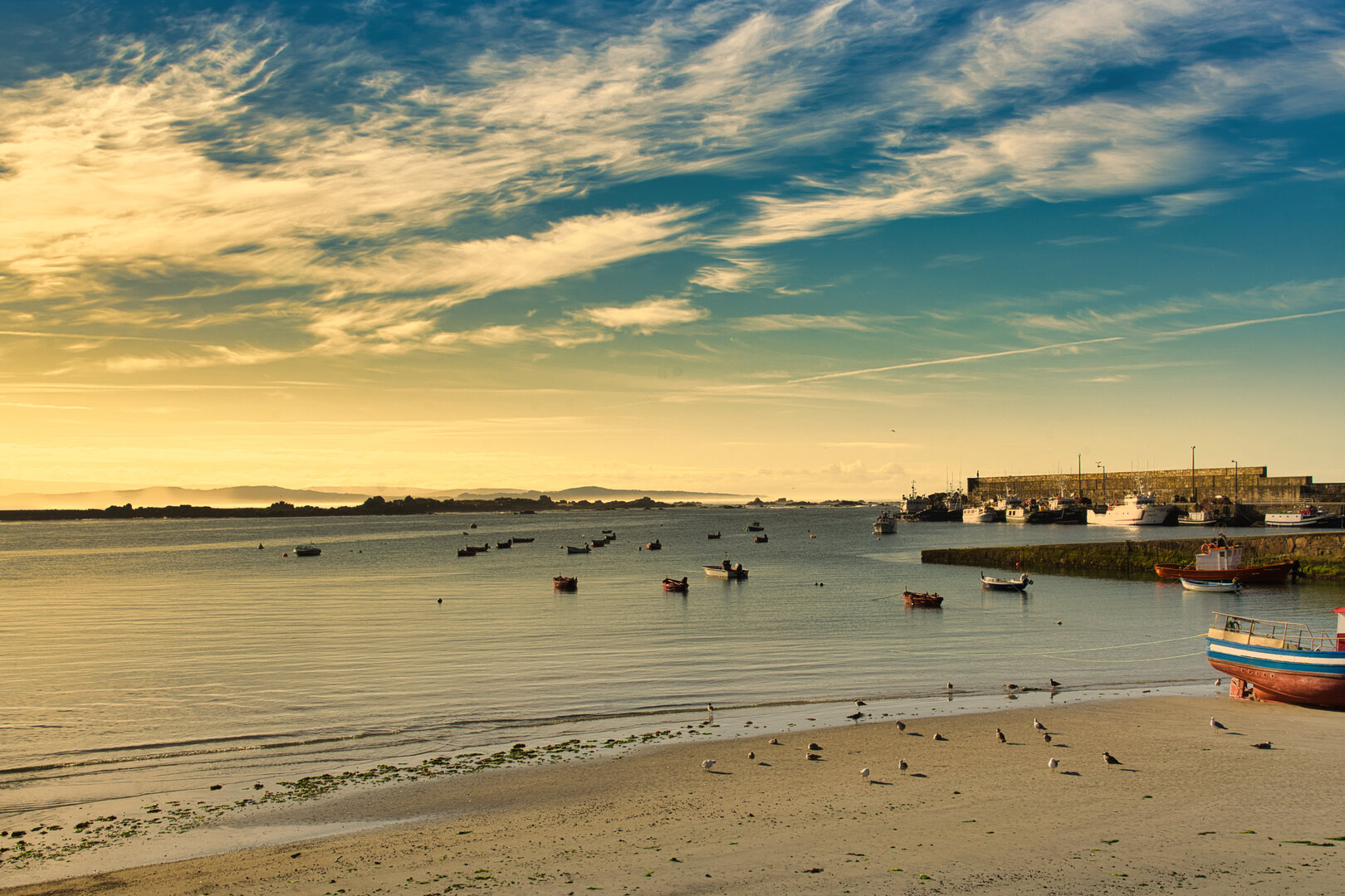 Vista panorámica del puerto de Aguiño al atardecer desde la terraza del ático, con barcas tradicionales, marea baja y cielo dorado