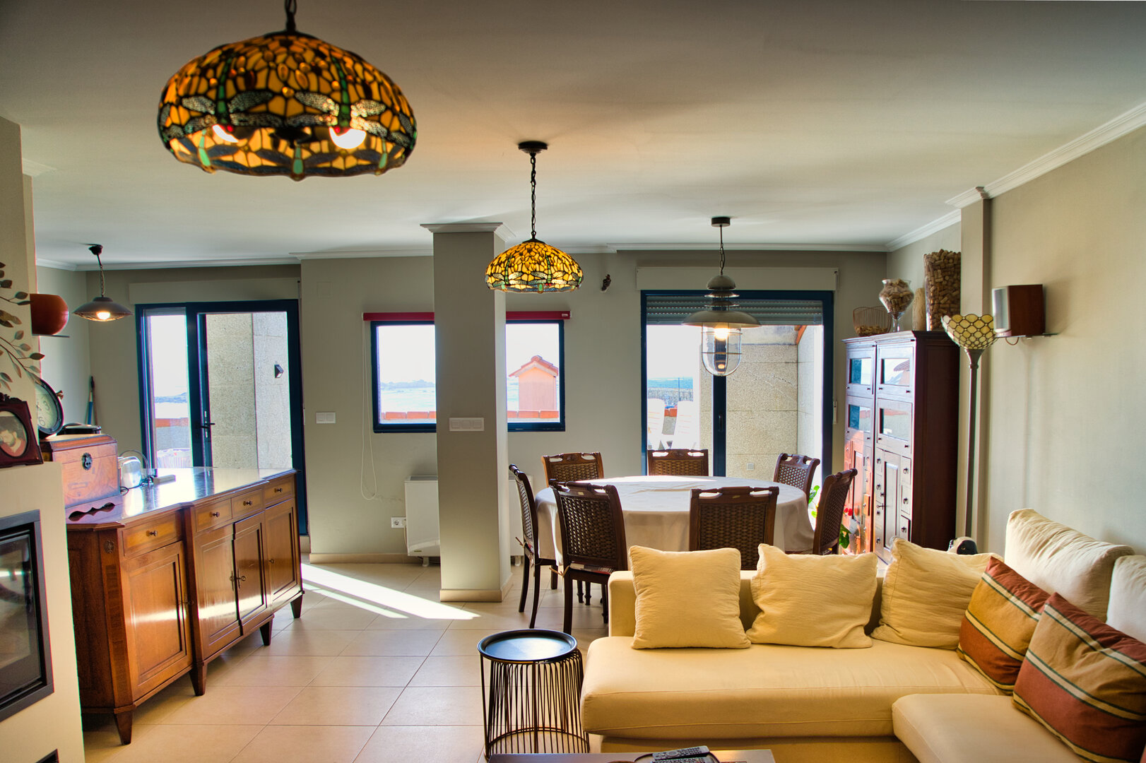 Salón-comedor del ático Illas Atlánticas con lámparas Tiffany, mobiliario de madera y grandes ventanales con vistas al puerto de Aguiño